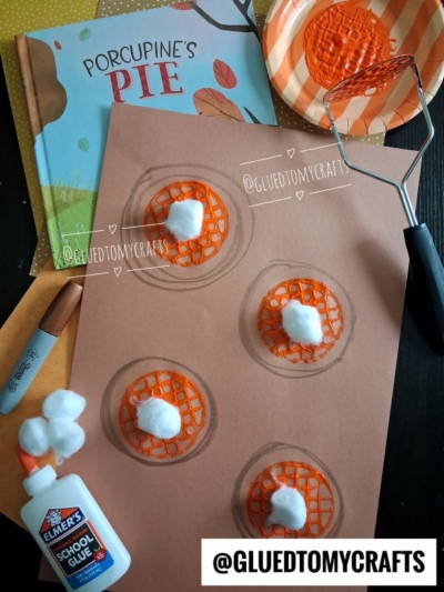 Brown paper with four orange pie crafts made from mesh and cotton balls, glue, a marker, a potato masher, a paper plate with orange paint for a fun potato masher stamped craft, and the Porcupines Pie book are arranged on a table.