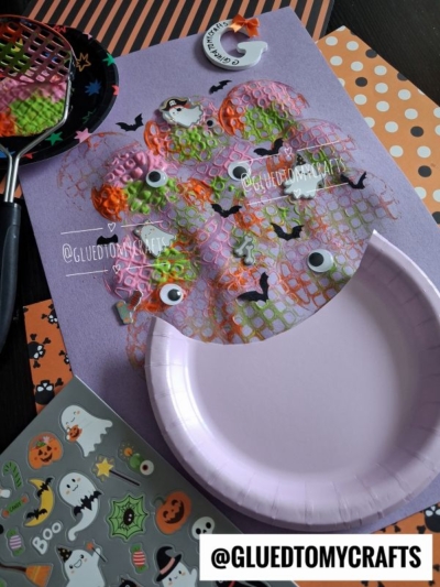 A Halloween craft project with bubble wrap painted in orange, green, and purple, googly eyes, bat stickers, and ghost stickers on purple paper. A potato masher stamped cauldron adds a whimsical touch alongside the purple plate and foam stickers, all set against festive background paper.