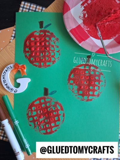 Green paper with three red apple prints, featuring brown stem and green leaf details, made using bubble wrap and paint. A red paint tray, roller, and other craft supplies—including a potato masher stamped apple—are visible nearby. @GLUEDTOMYCRAFTS is labeled on the image.