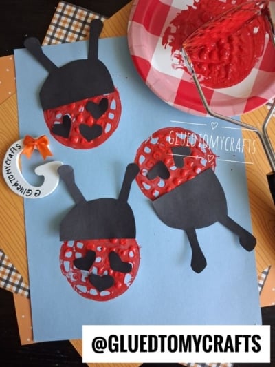 Three ladybugs made of red paint and black paper are arranged on blue paper as a craft. A plate with red paint, a whisk, and orange checks are visible. The text @GLUETOMYCRAFTS appears at the bottom.