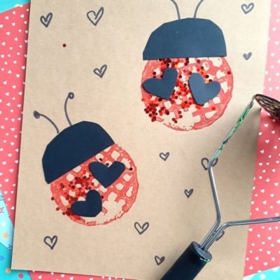 Two ladybugs made from red glitter and black paper hearts on brown paper, with a heart pattern background and a craft roller in the corner. The image includes the text @GLUEDTOMYCRAFTS.