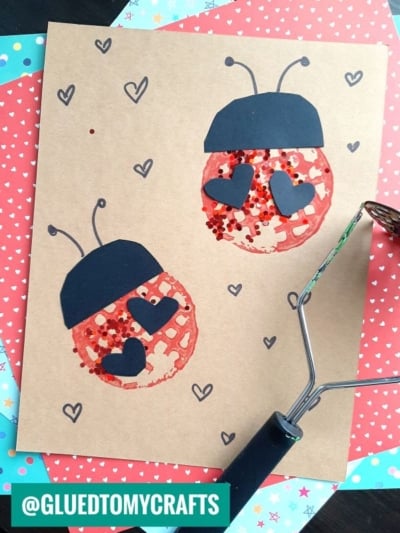 Two ladybugs made from red glitter and black paper hearts on brown paper, with a heart pattern background and a craft roller in the corner. The image includes the text @GLUEDTOMYCRAFTS.