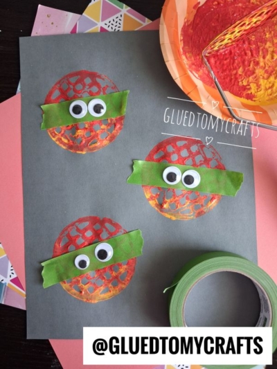 Three painted circles with green tape, googly eyes, and orange marks—created using a potato masher stamped craft technique—resemble Teenage Mutant Ninja Turtles on black paper. Tape, paper scraps, and a painted bowl are nearby. @GLUEDTOMYCRAFTS is labeled on the image.