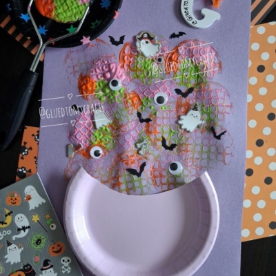 A Halloween craft features a paper plate with bubble wrap painted orange and pink, googly eyes, mini ghosts, and bat stickers. Surrounding are Halloween stickers and patterned papers, plus a playful potato masher stamped cauldron for an extra dash of spooky fun. @GLUEDTOMYCRAFTS watermark included.