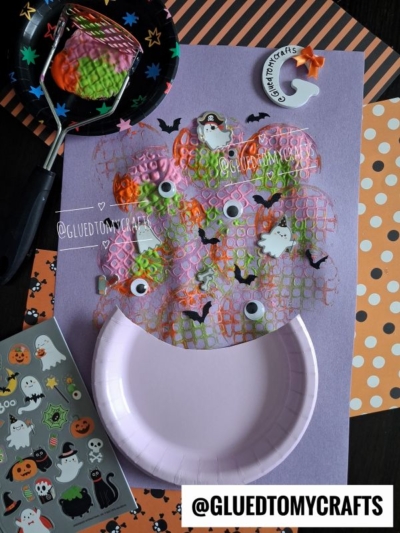 A Halloween craft features a paper plate with bubble wrap painted orange and pink, googly eyes, mini ghosts, and bat stickers. Surrounding are Halloween stickers and patterned papers, plus a playful potato masher stamped cauldron for an extra dash of spooky fun. @GLUEDTOMYCRAFTS watermark included.