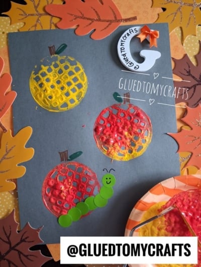 Childrens autumn craft with painted apple prints and a caterpillar on dark paper, surrounded by leaf cutouts. A small snail craft and GLUEDTOMYCRAFTS text appear on the art and in a white banner at the bottom.