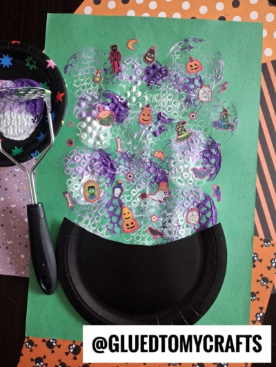 A Halloween craft featuring a black paper cauldron on green paper, foam bubble print, Halloween stickers, and festive scrapbook paper in the background. A black foam brush with Halloween patterns is on the left.