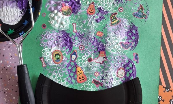 A Halloween craft featuring a black paper cauldron on green paper, foam bubble print, Halloween stickers, and festive scrapbook paper in the background. A black foam brush with Halloween patterns is on the left.