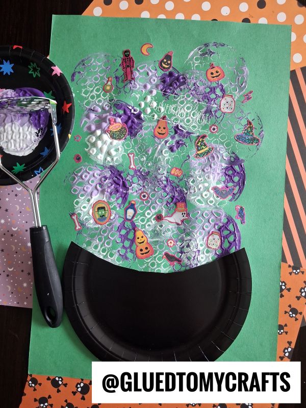A Halloween craft featuring a black paper cauldron on green paper, foam bubble print, Halloween stickers, and festive scrapbook paper in the background. A black foam brush with Halloween patterns is on the left.