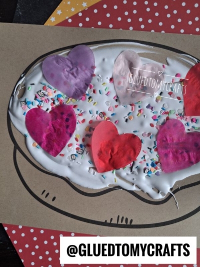 A paper craft of toast with white glue as frosting, topped with red and purple paper hearts and colorful sprinkles, on a polka dot background. Perfect for fans of puffy paint crafts, this whimsical piece adds a fun touch to any creative collection. @GLUETOMYCRAFTS is displayed at the bottom.