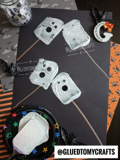 Children’s Halloween craft featuring ghost shapes made from white paint and popsicle sticks, with googly eyes on black paper, surrounded by festive Halloween decorations and accessories—this project combines classic spooky fun with a creative spatula stamped s’mores craft twist.