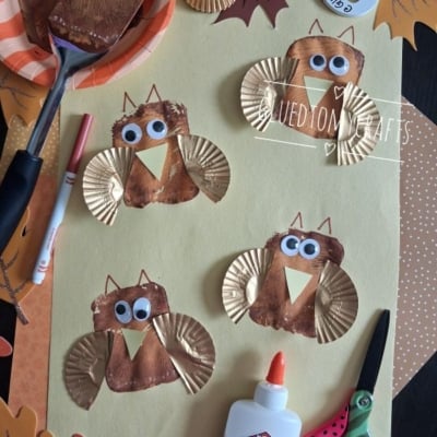 Four owl crafts made with gold cupcake liners, brown paint, googly eyes, and paper wings are arranged on yellow paper. Craft supplies like glue, scissors, and a paintbrush are also visible.