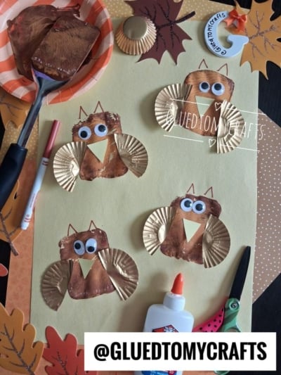 Four owl crafts made with gold cupcake liners, brown paint, googly eyes, and paper wings are arranged on yellow paper. Craft supplies like glue, scissors, and a paintbrush are also visible.