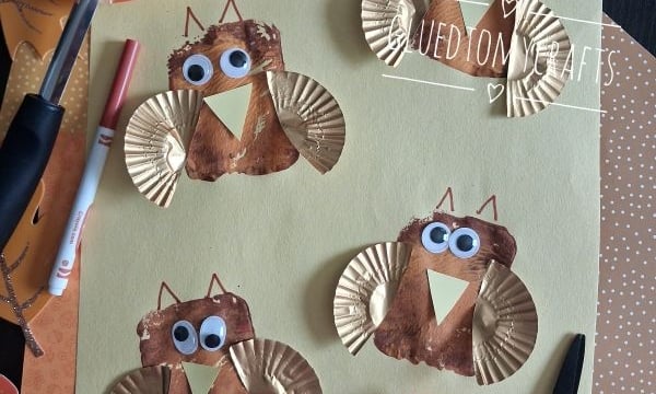 Four owl crafts made with gold cupcake liners, brown paint, googly eyes, and paper wings are arranged on yellow paper. Craft supplies like glue, scissors, and a paintbrush are also visible.
