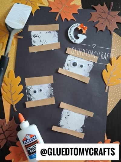 A Halloween craft features mummy faces on black paper with googly eyes, brown tape, and fake spider webs, surrounded by paper leaves, a spatula, and white glue. This spooky project takes inspiration from a spatula stamped smores craft for added creative fun. A tag reads @GLUEDTOMYCRAFTS.