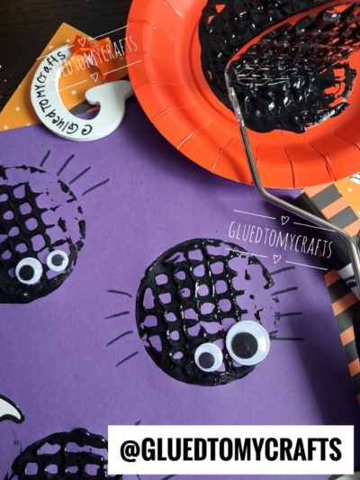 A purple paper with black spider prints made using a potato masher, each with googly eyes. Craft tools and an orange plate with black paint are also visible. @GLUEDTOMYCRAFTS is labeled on the image.