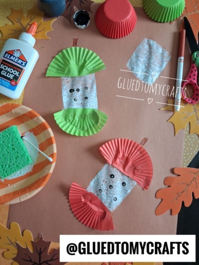 Two apple cores made from red and green cupcake liners, paper, and markers are displayed on a table with glue, a sponge, scissors, and autumn decorations—making this the perfect inspiration for FALL CRAFTS FOR KIDS. A watermark reads GLUEDTOMYCRAFTS.