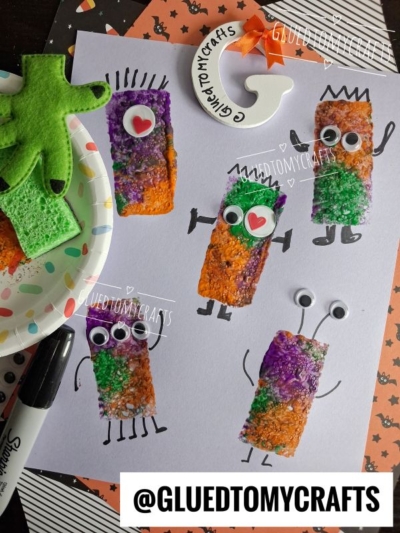 Four colorful sponge-stamped monsters with googly eyes and drawn limbs grace white paper, showcasing playful sponge painted monsters. A green sponge and marker are nearby, with Halloween-themed craft supplies surrounding the art. @GLUEDTOMYCRAFTS is displayed.