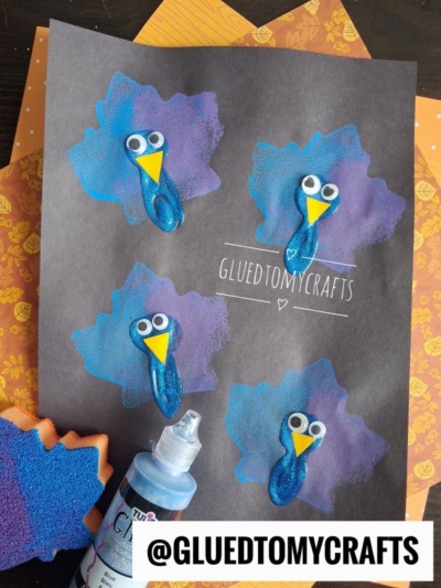 A black paper displays four blue and purple painted leaf shapes, each with a bird made from googly eyes, a yellow beak, and blue sparkle glue. Perfect for fall crafts for kids, bottles, a sponge, and autumn-patterned paper surround the artwork.