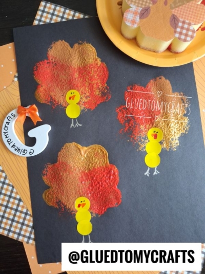 Children’s turkey crafts made with orange and red paint prints on black paper, decorated with yellow paper circles for bodies, and beaks and feet drawn on. Perfect as fall crafts for kids, these creations use simple craft supplies and are complemented by a pumpkin-themed plate nearby.