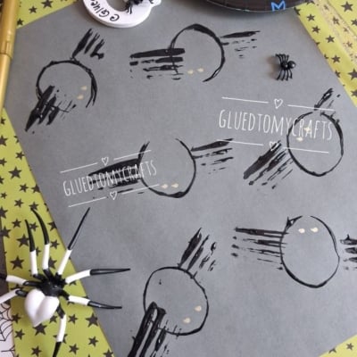 Completed toilet paper roll and fork painted spider craft on gray cardstock, featuring six black stamped spiders with gold eyes. A large realistic toy spider with a white abdomen and a small black plastic spider add spooky Halloween decor, set against lime-green star paper and spiderweb-patterned paper.