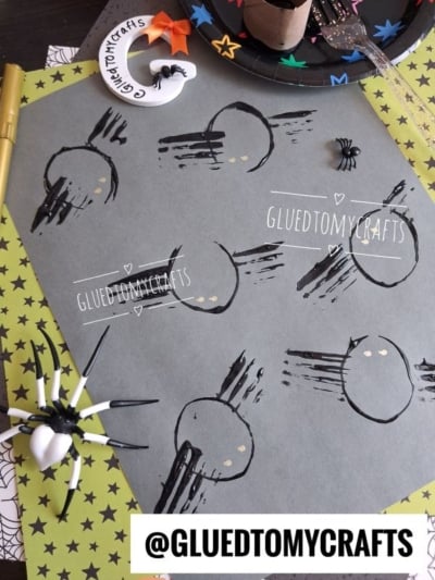 Completed toilet paper roll and fork painted spider craft on gray cardstock, featuring six black stamped spiders with gold eyes. A large realistic toy spider with a white abdomen and a small black plastic spider add spooky Halloween decor, set against lime-green star paper and spiderweb-patterned paper.