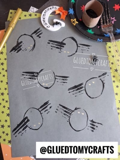 Flat-lay of a Halloween kids' craft showing six black spiders stamped on gray cardstock, made with a toilet paper roll for round bodies, a plastic fork for legs, and gold dots for eyes. Crafting tools surround the paper, including a gold marker, a decorated wooden letter "G," and a star-patterned paper plate holding the cardboard tube stamp and a glitter fork.