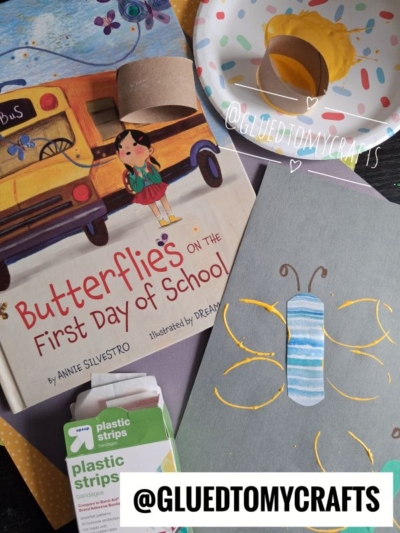 A childrens book, craft supplies, a painted butterfly made from a toilet paper roll, yellow paint on a plate, and the Instagram handle @GLUEDTOMYCRAFTS are shown on a table.