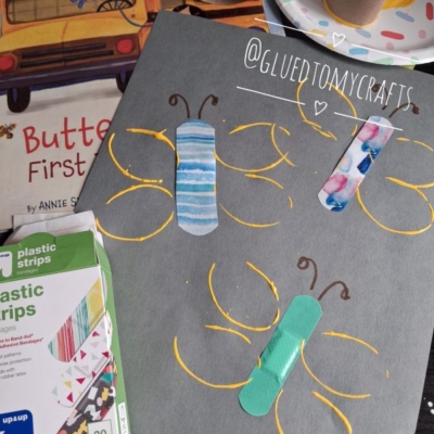A gray paper with butterflies made from colorful bandages and yellow paint. Art supplies and a butterfly book are nearby. @GLUEDTOMYCRAFTS is written on the photo.
