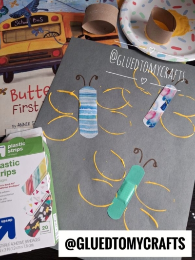 A gray paper with butterflies made from colorful bandages and yellow paint. Art supplies and a butterfly book are nearby. @GLUEDTOMYCRAFTS is written on the photo.