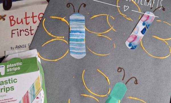 A gray paper with butterflies made from colorful bandages and yellow paint. Art supplies and a butterfly book are nearby. @GLUEDTOMYCRAFTS is written on the photo.