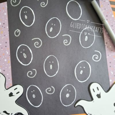 A black sheet with white ghost faces drawn on it, surrounded by Halloween-themed papers, two ghost cutouts, and a marker. The handle @GLUEDTOMYCRAFTS is visible at the bottom.