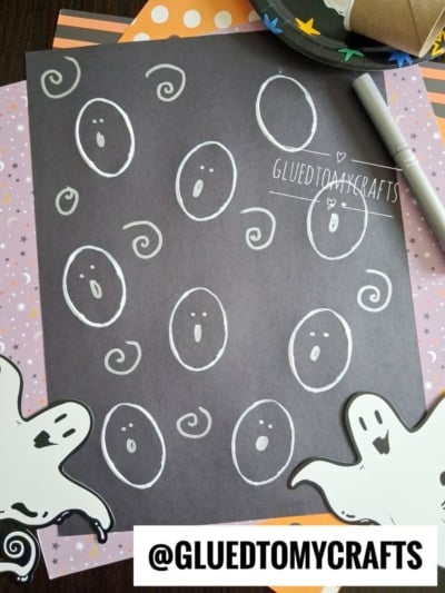 A black sheet with white ghost faces drawn on it, surrounded by Halloween-themed papers, two ghost cutouts, and a marker. The handle @GLUEDTOMYCRAFTS is visible at the bottom.