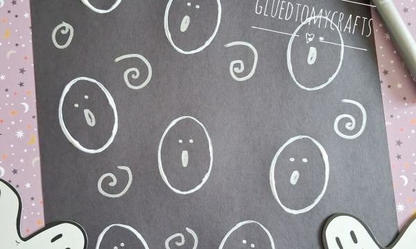 A black sheet with white ghost faces drawn on it, surrounded by Halloween-themed papers, two ghost cutouts, and a marker. The handle @GLUEDTOMYCRAFTS is visible at the bottom.