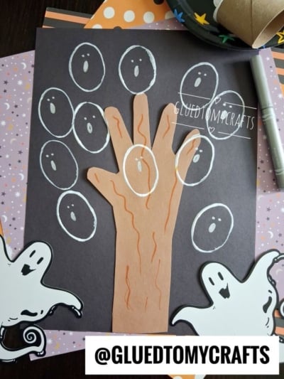 A Halloween craft with a hand-shaped cutout on black paper, surrounded by white-drawn ghosts and two ghost cutouts. A pen and orange papers are in the background. @GLUEDTOMYCRAFTS is labeled at the bottom.