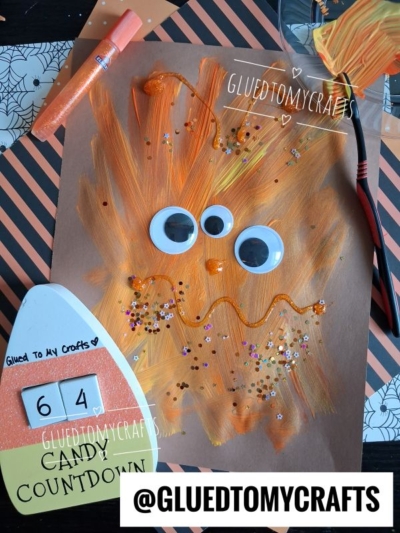 Children’s Halloween monster craft on brown paper featuring a spiky orange toothbrush-painted creature with three googly eyes, glitter details, and craft supplies arranged around it.