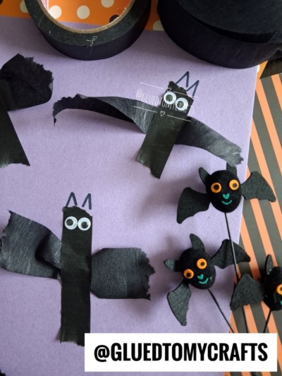 Two handmade bat crafts made from black crepe paper, popsicle sticks, googly eyes, and paper, displayed on purple and orange-striped backgrounds. A watermark and the text @GLUEDTOMYCRAFTS appear on the image.