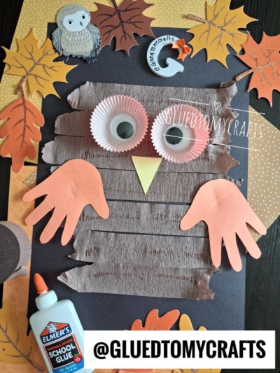 A handmade owl craft on black paper with cupcake liners for eyes, handprint wings, a yellow beak, paper leaves, and a glue bottle—perfect inspiration for FALL CRAFTS FOR KIDS. The handle @GLUEDTOMYCRAFTS is at the bottom.