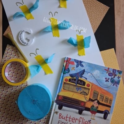 A butterfly craft project with blue and yellow paper butterflies, crafting materials, and the book Butterflies on the First Day of School displayed on patterned paper.