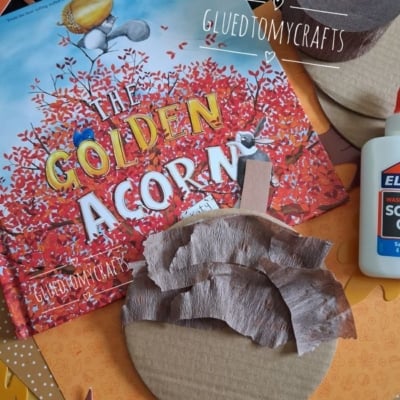 Childrens craft supplies with a book titled The Golden Acorn, glue, and cardboard circles partly covered with brown tissue paper, surrounded by colorful paper leaves. @GLUEDTOMYCRAFTS is written on the image.