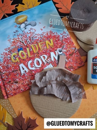 Childrens craft supplies with a book titled The Golden Acorn, glue, and cardboard circles partly covered with brown tissue paper, surrounded by colorful paper leaves. @GLUEDTOMYCRAFTS is written on the image.