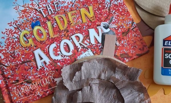 Childrens craft supplies with a book titled The Golden Acorn, glue, and cardboard circles partly covered with brown tissue paper, surrounded by colorful paper leaves. @GLUEDTOMYCRAFTS is written on the image.