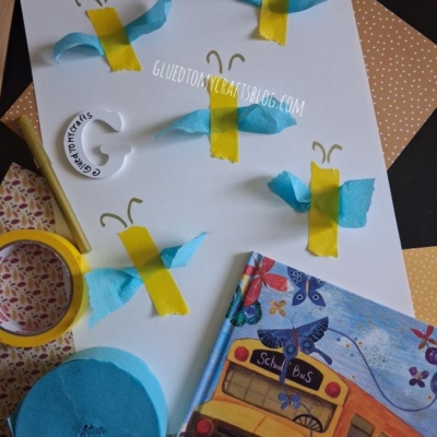 A craft project with blue and yellow tissue paper butterflies on white paper, surrounded by yellow tape, blue tissue, a round white label, and a childrens book featuring a school bus. @GLUEDTOMYCRAFTS watermark is visible.