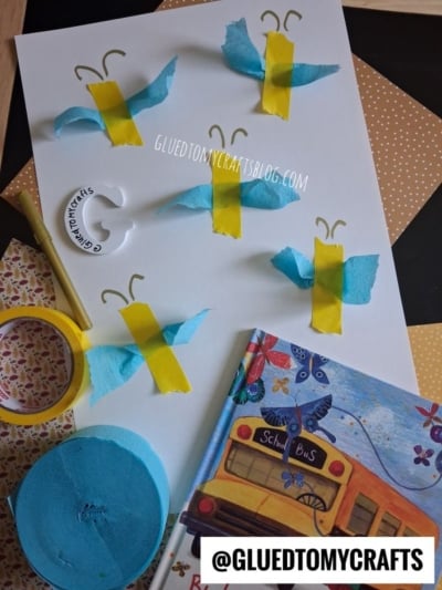 A craft project with blue and yellow tissue paper butterflies on white paper, surrounded by yellow tape, blue tissue, a round white label, and a childrens book featuring a school bus. @GLUEDTOMYCRAFTS watermark is visible.