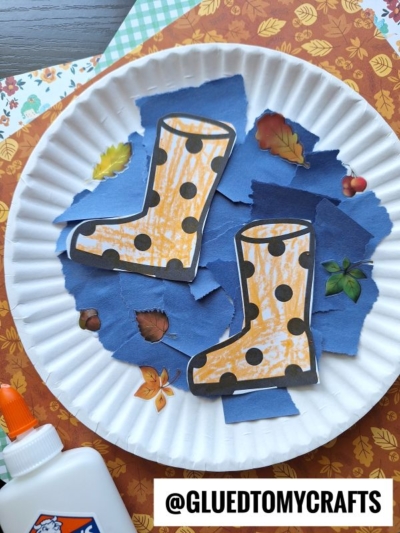 A paper plate with a torn paper rain puddle made from blue paper, decorated with leaf stickers and two orange polka-dot rain boots cutouts. A glue bottle is nearby and patterned paper is in the background. @GLUEDTOMYCRAFTS text is at the bottom.