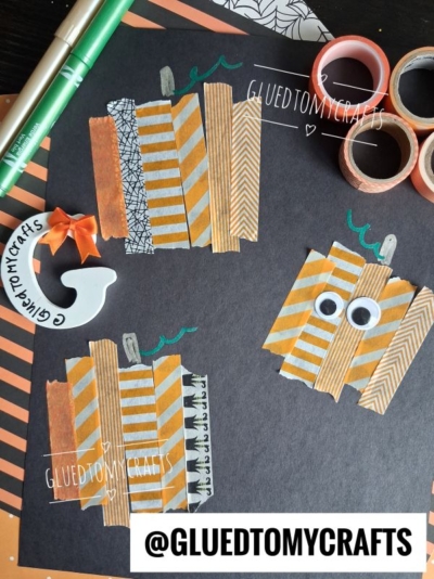 Two pumpkins made from orange washi tape strips are on black paper, with one pumpkin sporting googly eyes. Perfect for FALL CRAFTS FOR KIDS, this scene features crafting supplies nearby, including washi tape rolls, markers, and a tag with a ribbon.