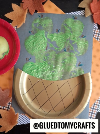 A craft project on gray paper featuring an apple stamped basket, with green apple-print stamps sitting above a gold paper plate cut in half to resemble a pie crust.