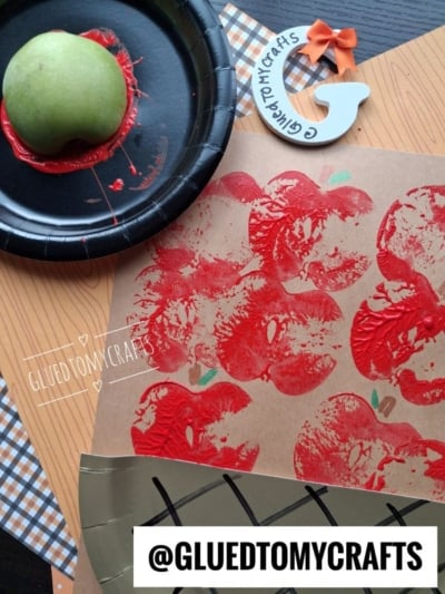 An apple dipped in red paint on a plate, apple prints on brown paper, plaid paper, an orange bow, and @GLUEDTOMYCRAFTS text label are shown on a crafting table.
