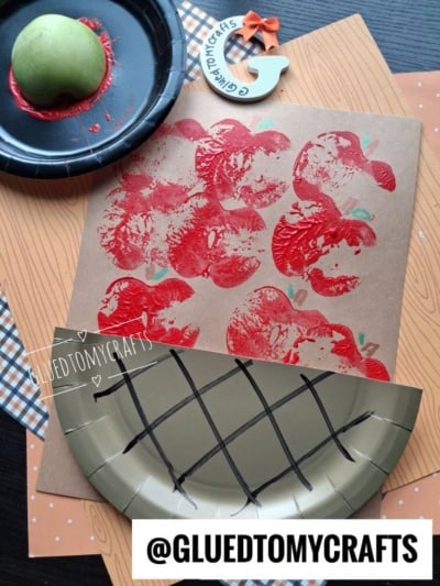 Apple prints made with a real apple and red paint on brown paper, a green apple, and gold paper plate with black marks arranged on a wooden table with craft supplies. @GLUEDTOMYCRAFTS label at the bottom.