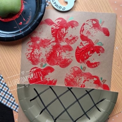 A craft project shows red apples stamped on paper, made using half an apple and red paint. Theres a paper plate designed to look like a basket and a @GLUEDTOMYCRAFTS label at the bottom.